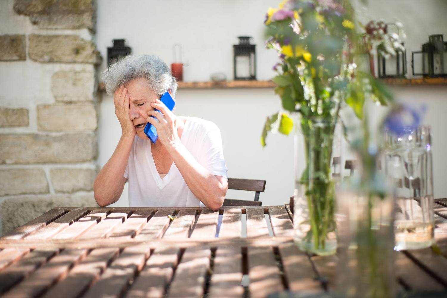 An older woman at an outdoor table on the phone, holding her jaw in discomfort